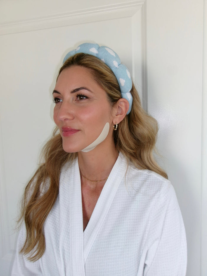 Woman wearing a white robe and a soft blue headband with white clouds, applying Jawline Sculpting Patches | Seald™ Wellness to enhance her skincare routine and facial contour.