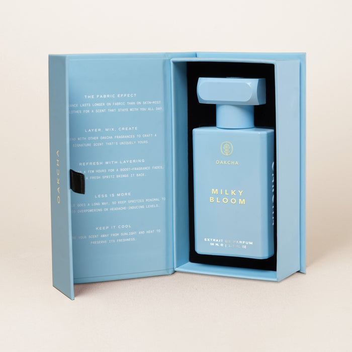 Front view of Milky Bloom perfume bottle in open pale blue gift box showing rectangular bottle with gold lettering on beige background