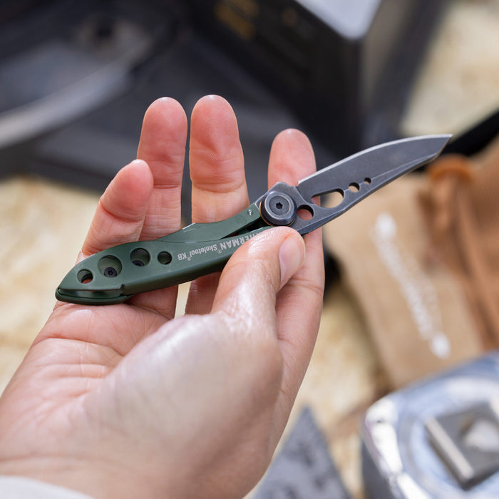 Hand holding an OD green Leatherman Skeletool KB folding knife with open blade.