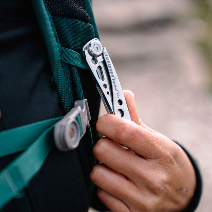 Hand clipping a stainless steel Skeletool KBX pocket knife onto a backpack strap.