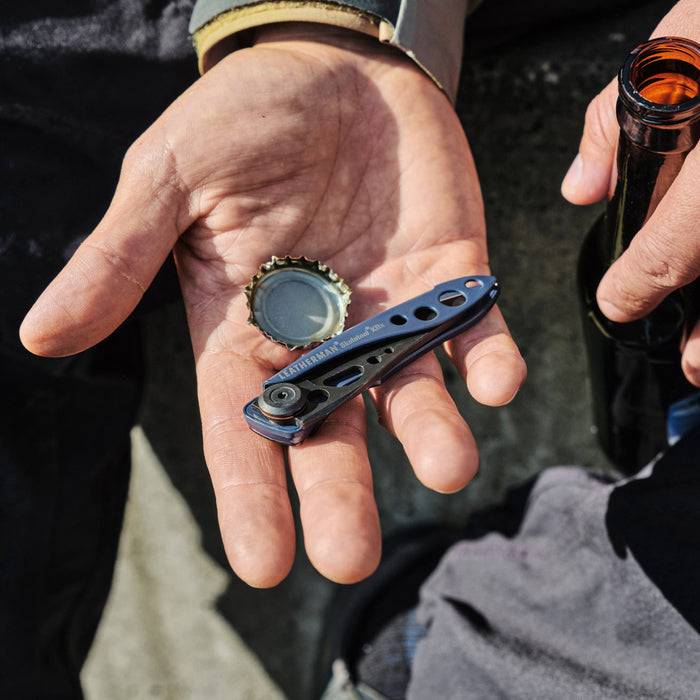 Hand holding a blue Skeletool KBX and bottle cap next to a person gripping an open glass bottle.