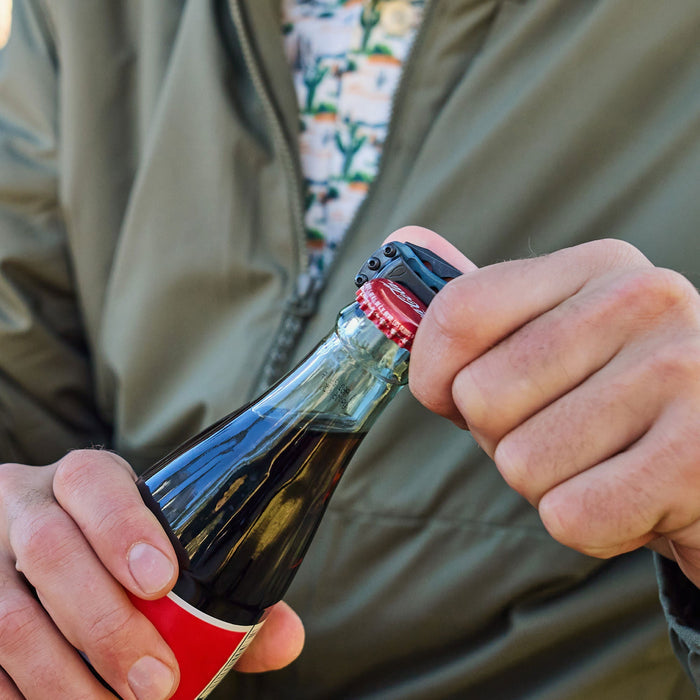 Hand using a Skeletool KBX to pry off the cap of a glass soda bottle.