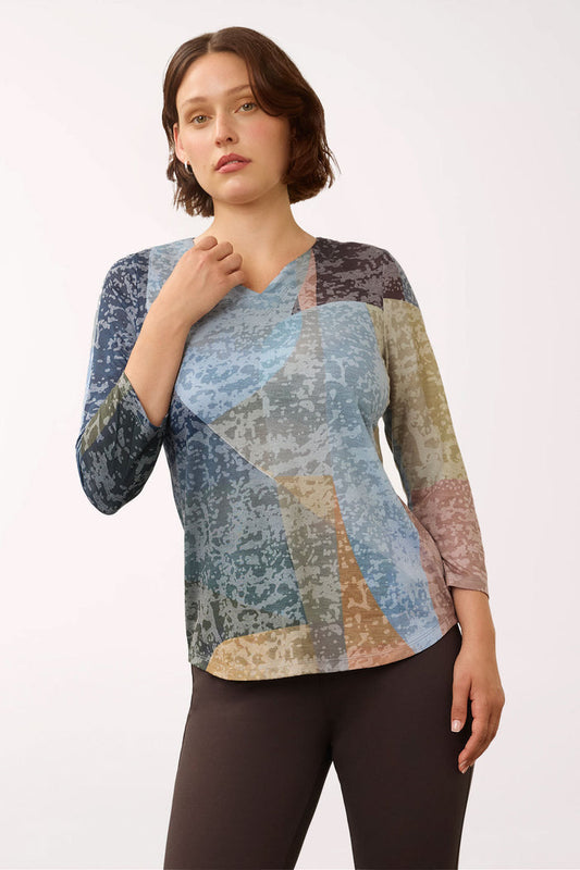 A woman modeling a long-sleeve V-neck top with a multicolor abstract patchwork print in shades of blue, beige, brown, and gray, paired with dark brown leggings, against a plain white studio background.